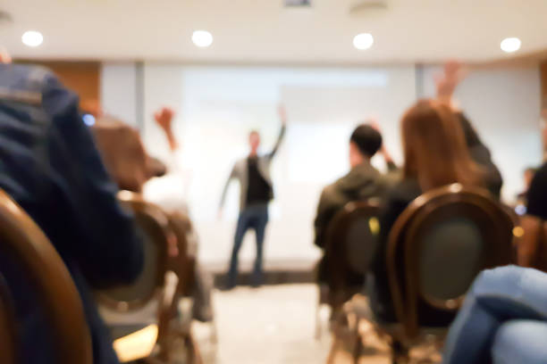 blurred group of people meeting in motivation seminar event at convention hall, speaker raising hand up and audience action follow , cheerful concept